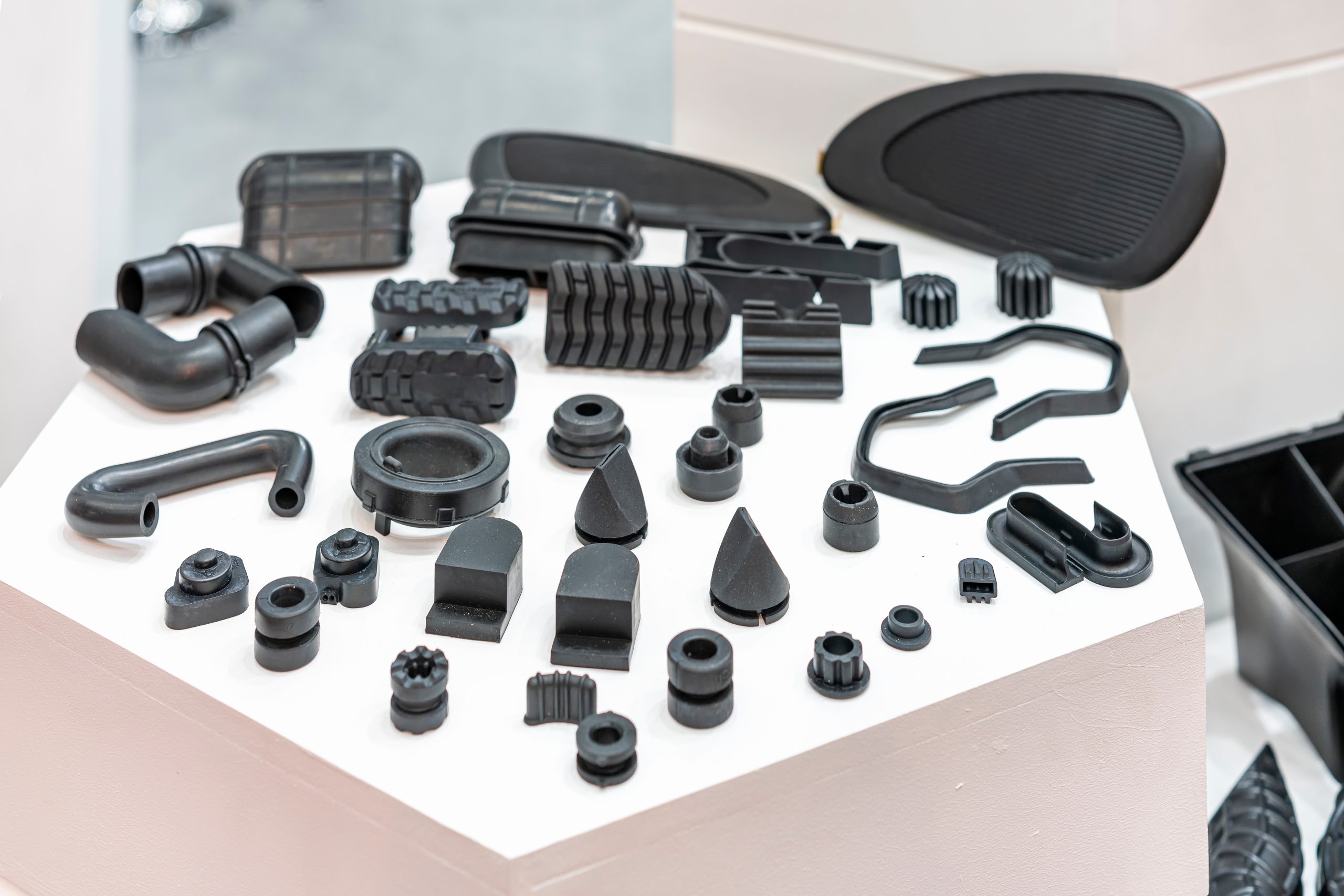 A wide selection of matte black molded industrial rubber and plastic parts, including elbow pipes, grommets, gaskets, and textured pads, neatly arranged on a white display pedestal.